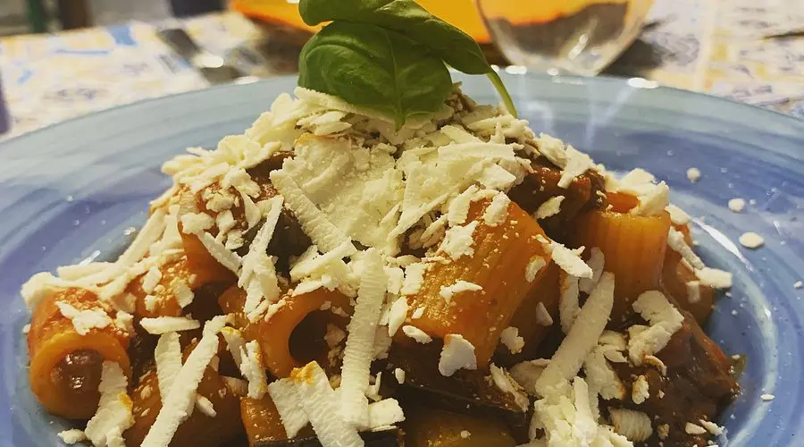 Tradizionale piatto di pasta alla Norma con melanzane fritte, ricotta salata grattugiata e basilico fresco, servito con salsa di pomodoro arricchita di aglio e olio d'oliva. Un piatto tipico della cucina siciliana, ricco di sapori autentici e mediterranei.