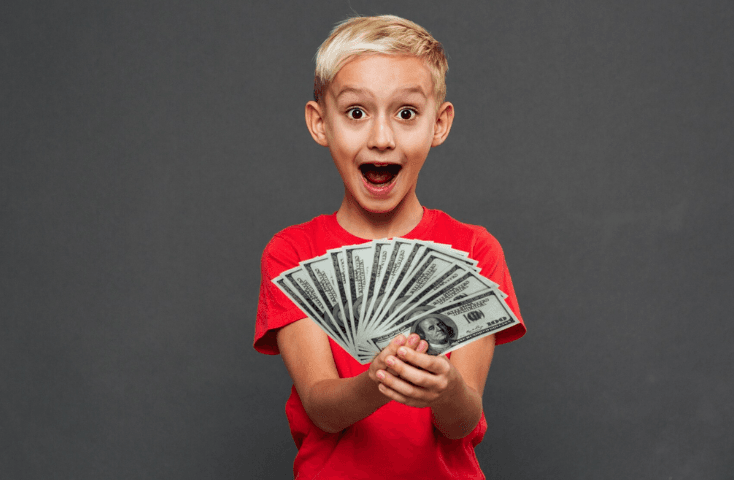 Bambino biondo felice e sorpreso con maglietta rossa tiene in mano soldi banconote come ventaglio su fondo grigio, come si dice bocca in inglese