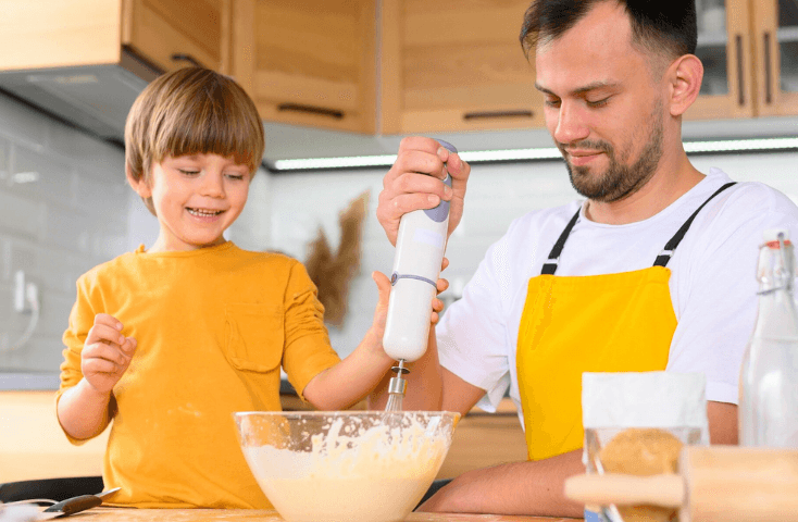 Papà e figlio che preparano un impasto col mixer, felici coi grembiuli gialli, cucinare insieme in cucina, vista frontale, quattro ricette dolci da preparare con i bambini