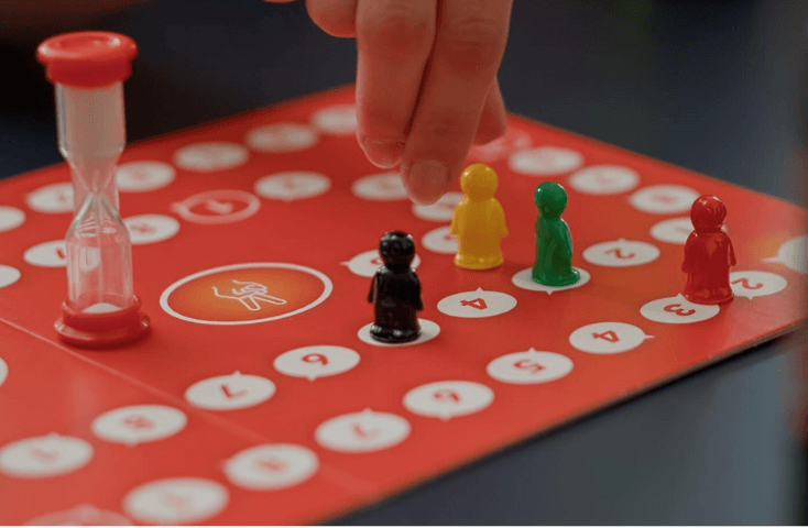 Mano sopra un tabellone da gioco rosso con pedine a forma di omini giallo, verde, nero e una clessidra, dieci giochi da tavolo da fare coi bambini in inglese