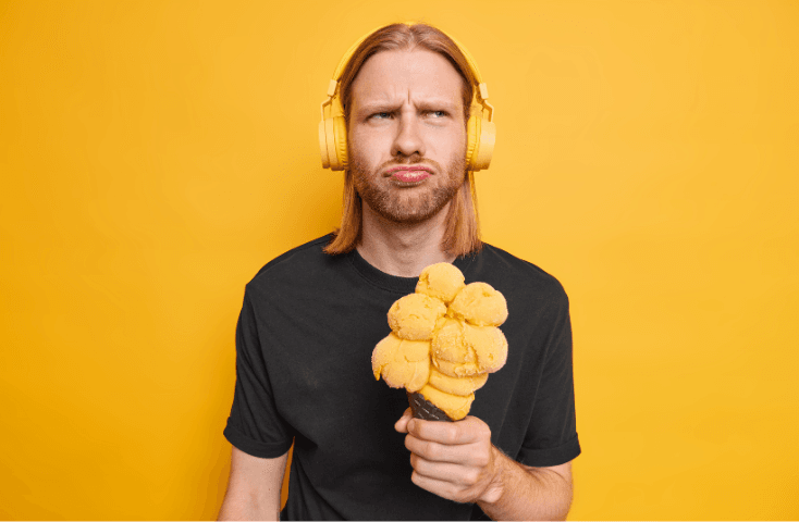 Ragazzo giornalista perplesso ascolta musica con cuffie stereo con maglia nera su parete gialla tiene in mano un microfono cono gelato, vista frontale, giochi di parole in inglese