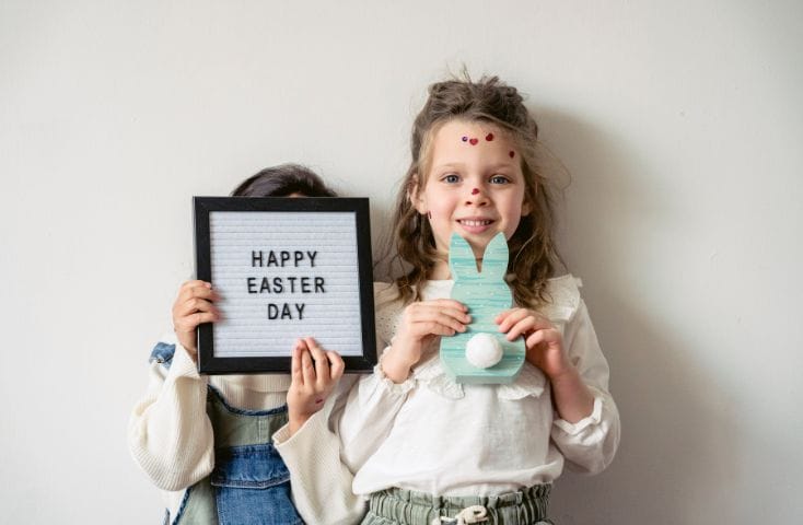 Bbambina con in mano un coniglietto di cartoncino e bambini con un cartello con scritto Happy Easter davnti l viso, come si dice Buona Pasqua in inglese