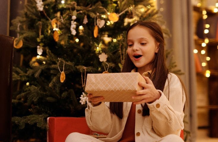 Bambina davanti all'albero di Natale con in mano un pacchetto regalo e con la faccia piena di stupore