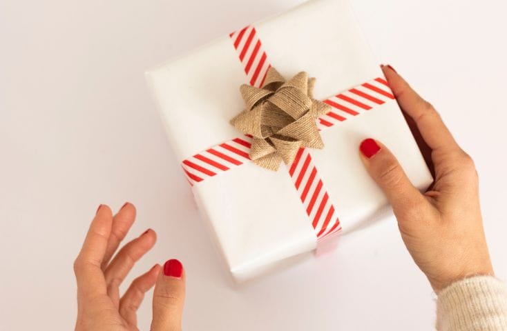 Mani che porgono un regalo impacchettato con la carta bianca e un nastro bianco e rosso, con coccarda dorata