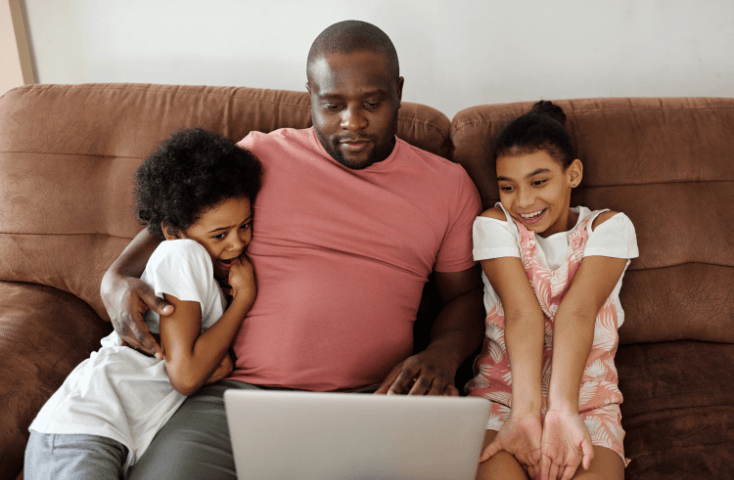 papà che guarda i cartoni sul divano con le due figlie davandi a un portatile